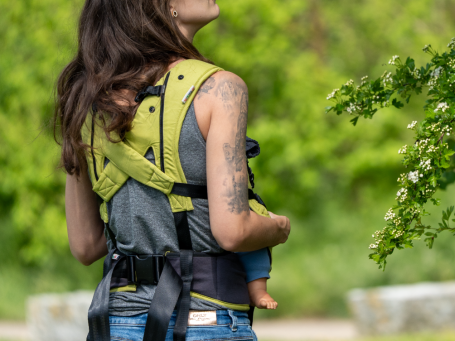 Frau mit Tätowierung trägt ein Baby in einer Tragehilfe, man sieht den Rücken und die besondere Gurtführung der Trage